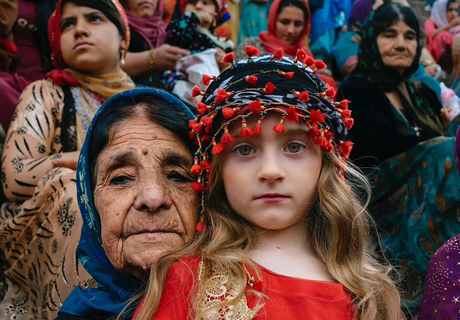 A grandmother and her grandchild watch the camera, as if deep in thought. The grandmother has deep set wrinkles, wears a blue headscarf, and leans close to the girl. The girl is wearing a red outfit with lace detailing. A grandmother and her grandchild watch the camera, as if deep in thought. The grandmother has deep set wrinkles, wears a blue headscarf, and leans close to the girl. The girl is wearing a red outfit with lace detailing.
