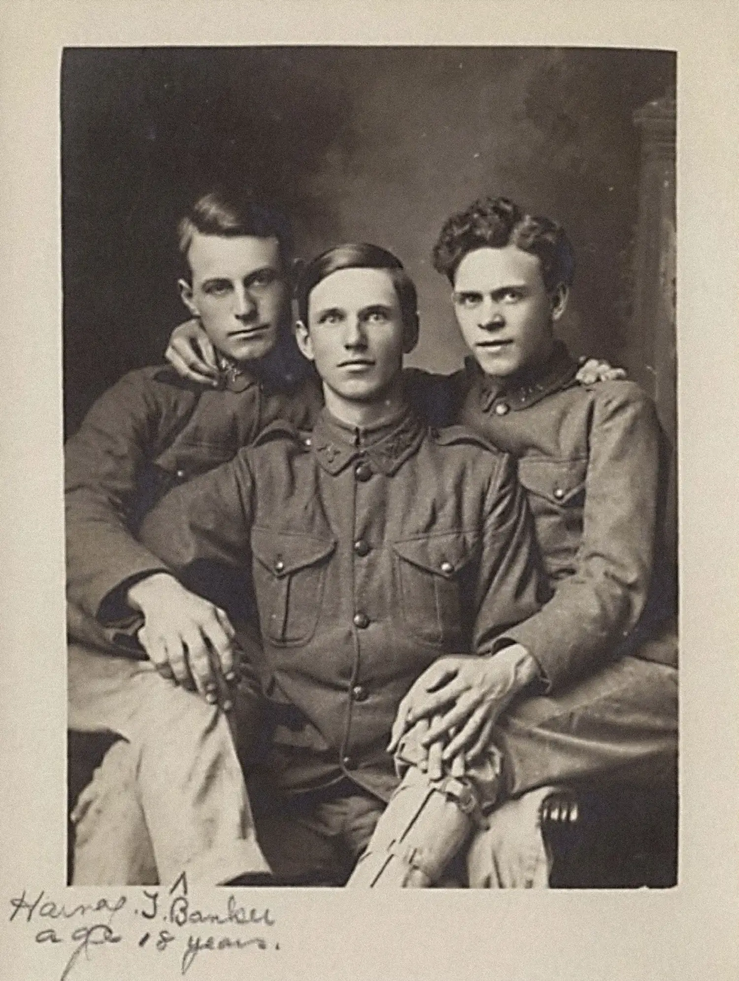 Three young men in WW1-era military uniform are seated together in an embrace. The man in the center is holding the hands of the two men seated on each side, and the outer two men each have a hand draped around the other’s shoulder. Three young men in WW1-era military uniform are seated together in an embrace. The man in the center is holding the hands of the two men seated on each side, and the outer two men each have a hand draped around the other’s shoulder.