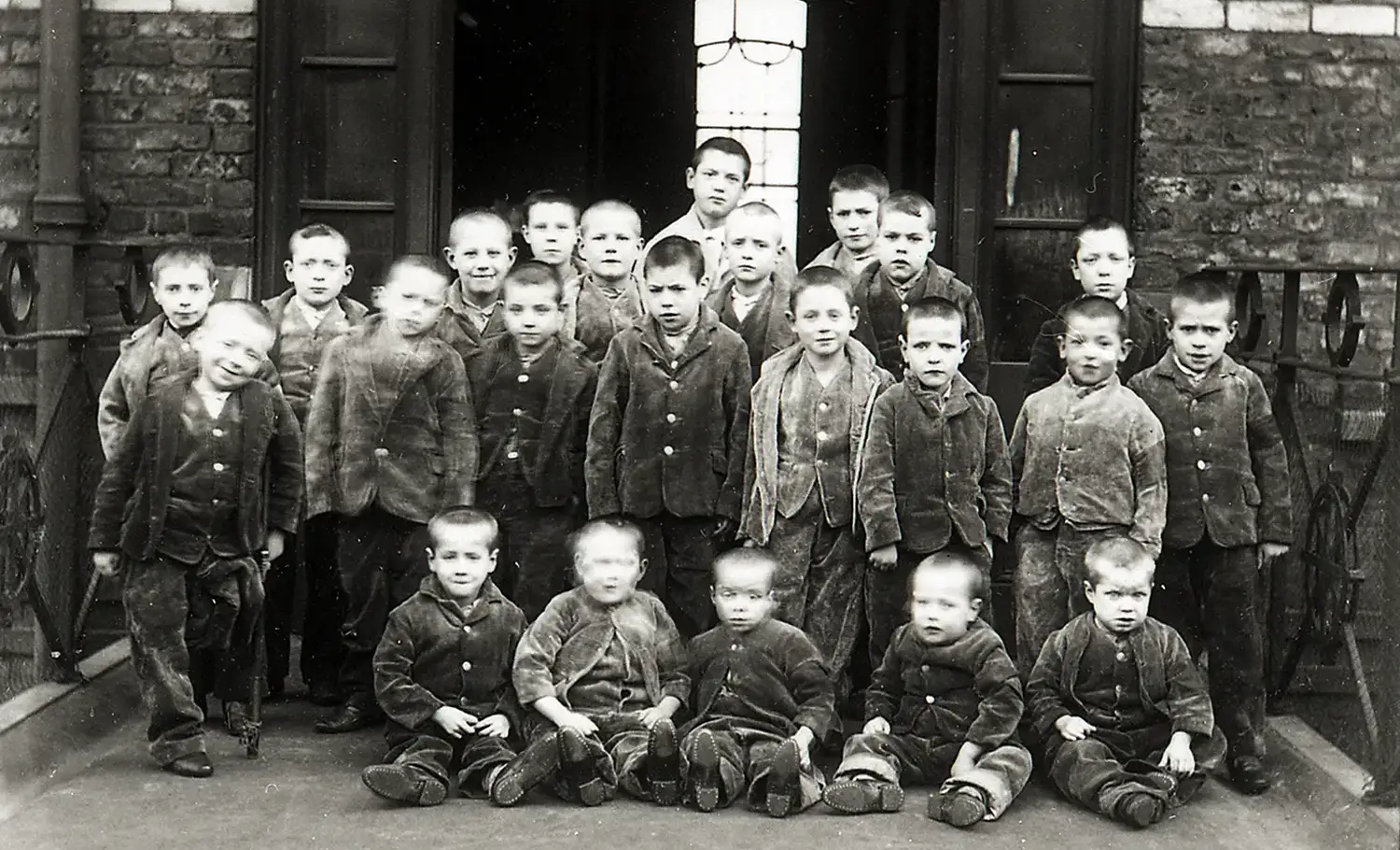 Twenty-three boys ranging from toddlers to about twelve years old are arranged for this group photograph. They all have their heads shaved and are wearing identical work clothes. The youngest sit in front, and one child appears to be missing a leg and using a cane. Twenty-three boys ranging from toddlers to about twelve years old are arranged for this group photograph. They all have their heads shaved and are wearing identical work clothes. The youngest sit in front, and one child appears to be missing a leg and using a cane.