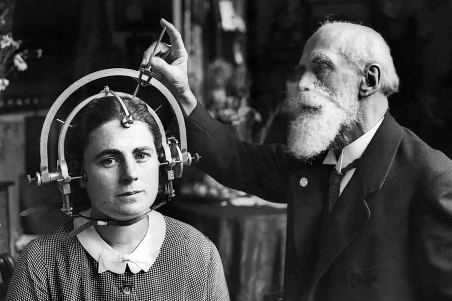 A woman sits with an elaborate circular measurement device strapped around her head. A much older bearded man adjusts the device while she stares into the camera. A woman sits with an elaborate circular measurement device strapped around her head. A much older bearded man adjusts the device while she stares into the camera.