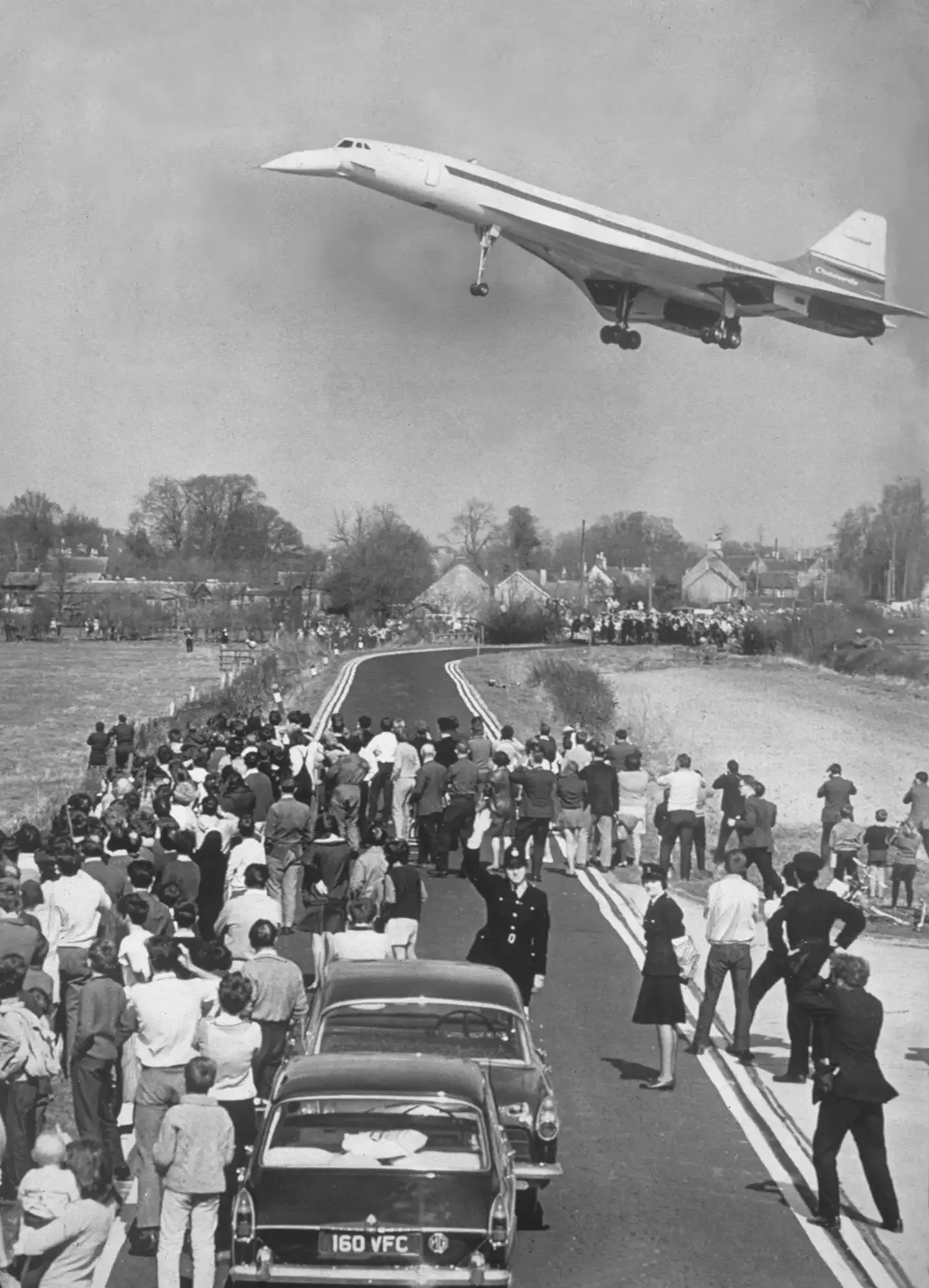 The Concorde aircraft flies low in the sky over a crowd of onlookers standing in the street below. The streamlined aircraft juxtaposes with the black and chrome mid-century cars stopped in the street. The Concorde aircraft flies low in the sky over a crowd of onlookers standing in the street below. The streamlined aircraft juxtaposes with the black and chrome mid-century cars stopped in the street.