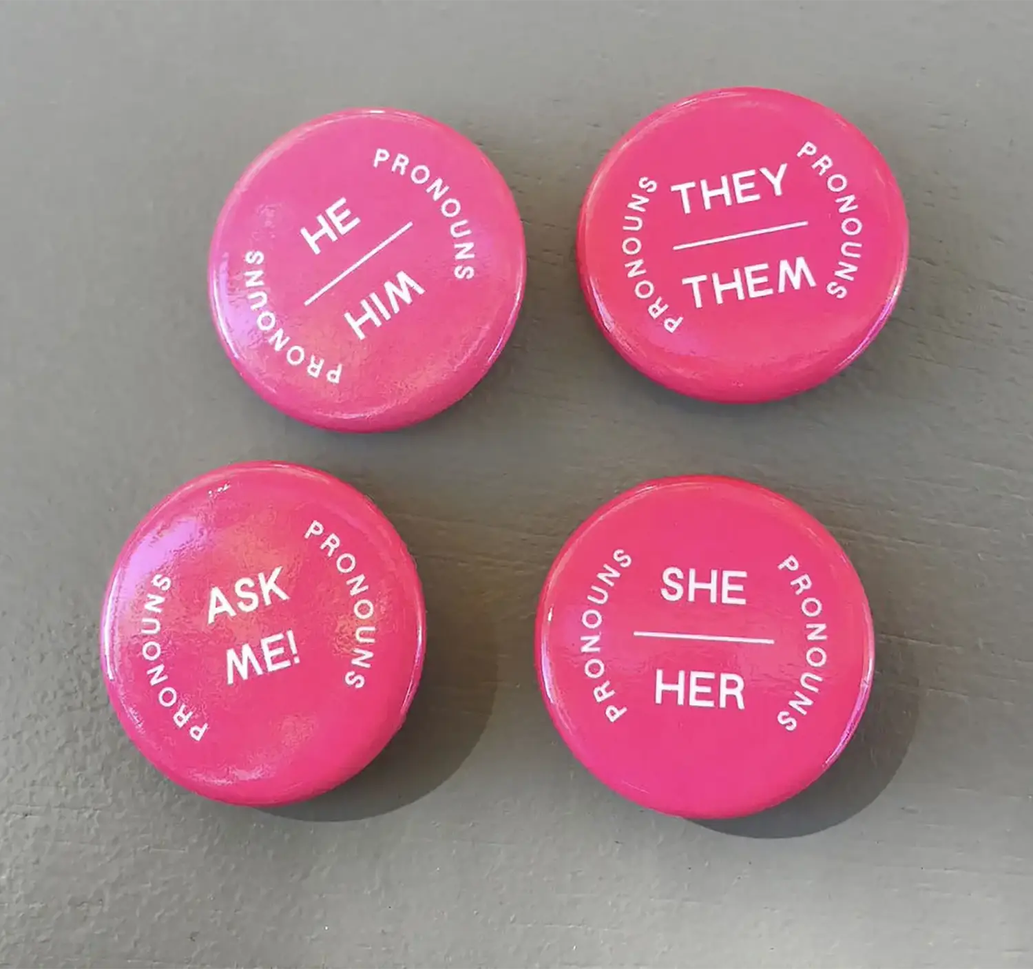 Four bright pink pronoun pins. Around the edge of each, it says “Pronouns,” in the center of each it says, respectively, “They/Them,” “She/Her,” “He/Him,” and “Ask Me!” Four bright pink pronoun pins. Around the edge of each, it says “Pronouns,” in the center of each it says, respectively, “They/Them,” “She/Her,” “He/Him,” and “Ask Me!”