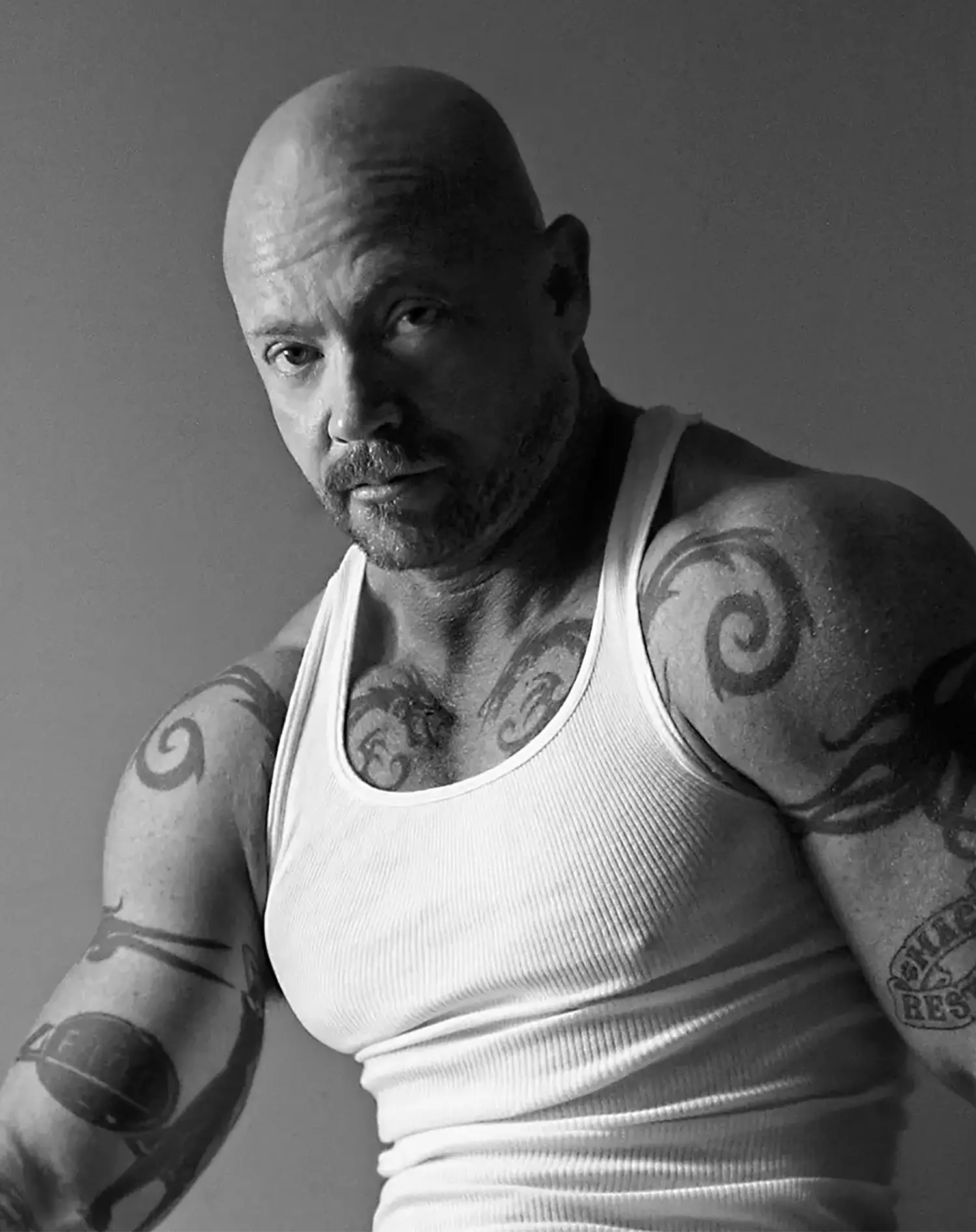 A studio portrait of a bald and bearded Buck Angel looking intently at the camera. He is wearing a white tank top that reveals arm and chest tattoos. A studio portrait of a bald and bearded Buck Angel looking intently at the camera. He is wearing a white tank top that reveals arm and chest tattoos.