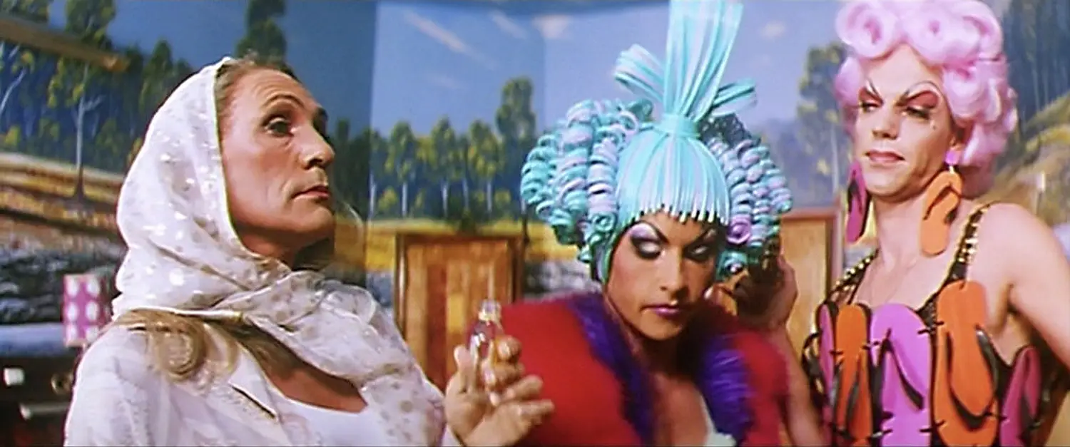 Movie still from The Adventures of Priscilla Queen of the Desert, showing the three main characters, one of which is trans and the other two who are dressed in colorful, flamboyant drag outfits. Movie still from The Adventures of Priscilla Queen of the Desert, showing the three main characters, one of which is trans and the other two who are dressed in colorful, flamboyant drag outfits.